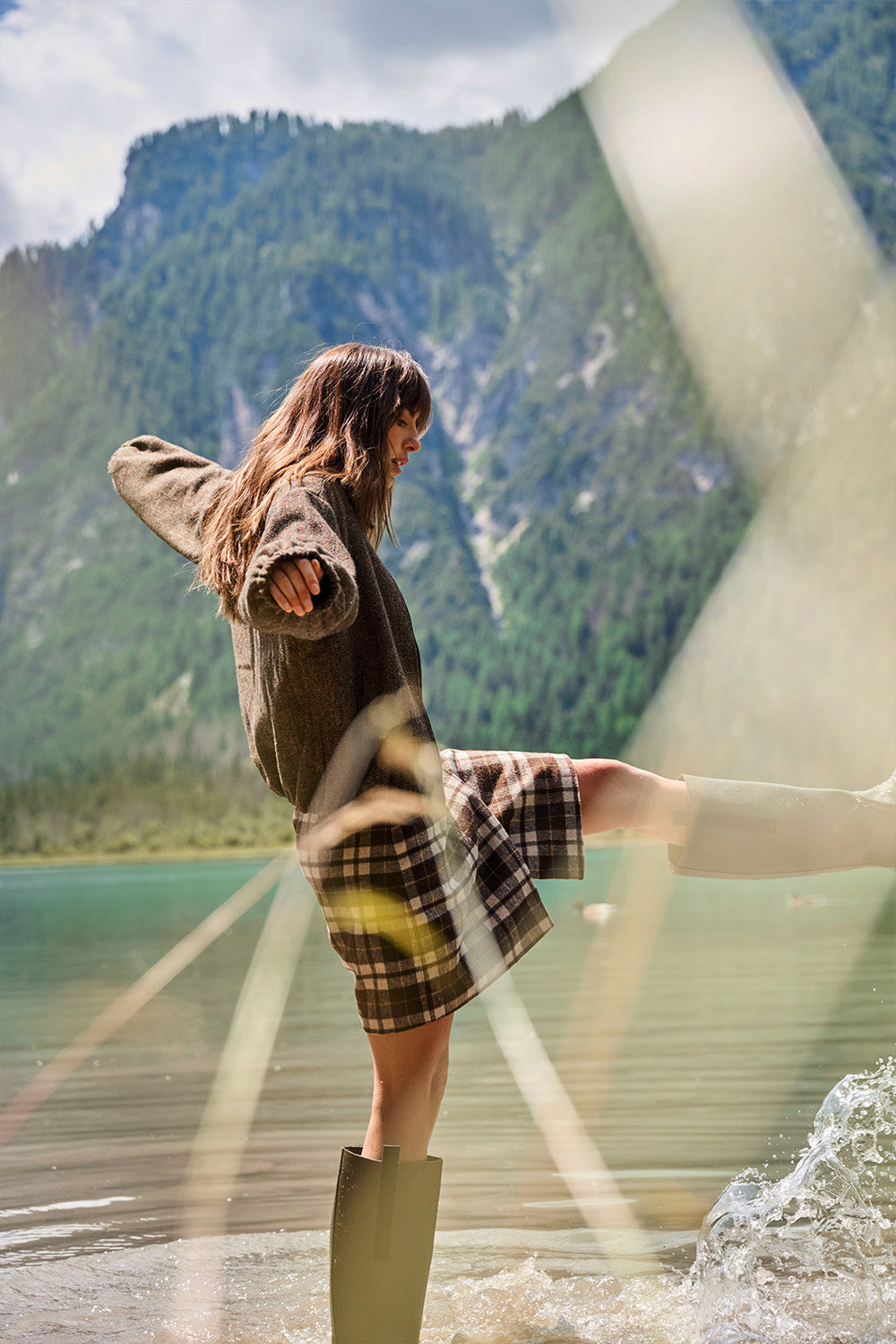 A woman stands in shallow water at a mountain lake, wearing a brown oversized sweater, a plaid skirt, and tall dark rain boots. The photo is taken through a reflective or textured surface with diagonal light streaks across the frame.