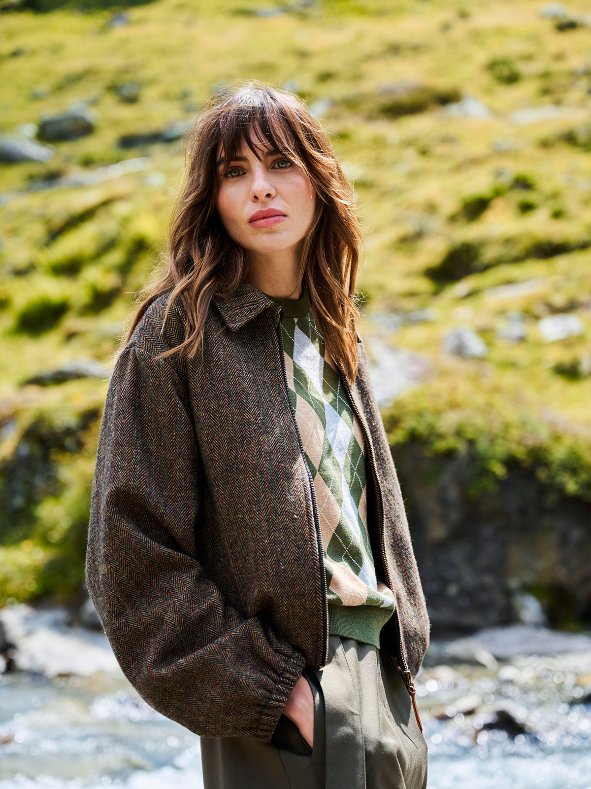 A woman stands outdoors in front of a rocky stream with a grassy hill in the background, wearing a brown textured zip-up jacket over a green and beige argyle sweater, paired with olive green trousers.