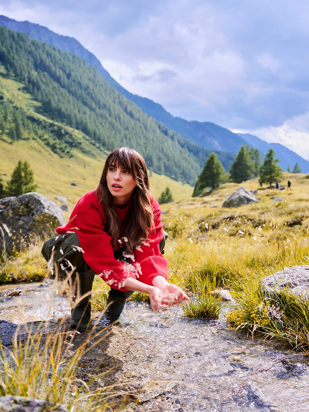 A woman in a red sweater and black pants crouches in a rocky mountain stream, reaching toward the water, with green hills and blue mountains in the background.