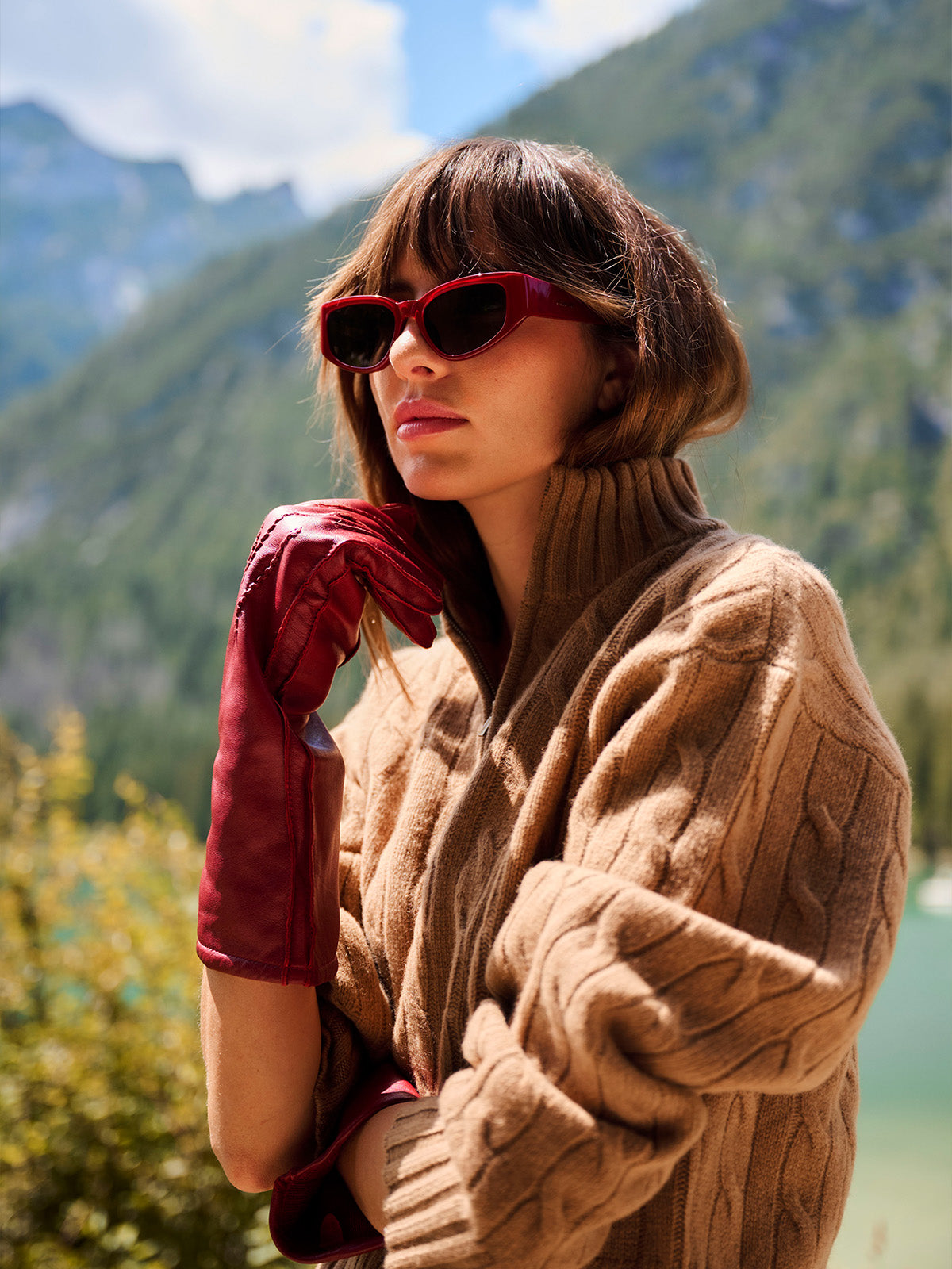 A woman wearing red cat-eye sunglasses, a brown cable-knit sweater, and red leather gloves, posed outdoors against a scenic mountain and lake background.